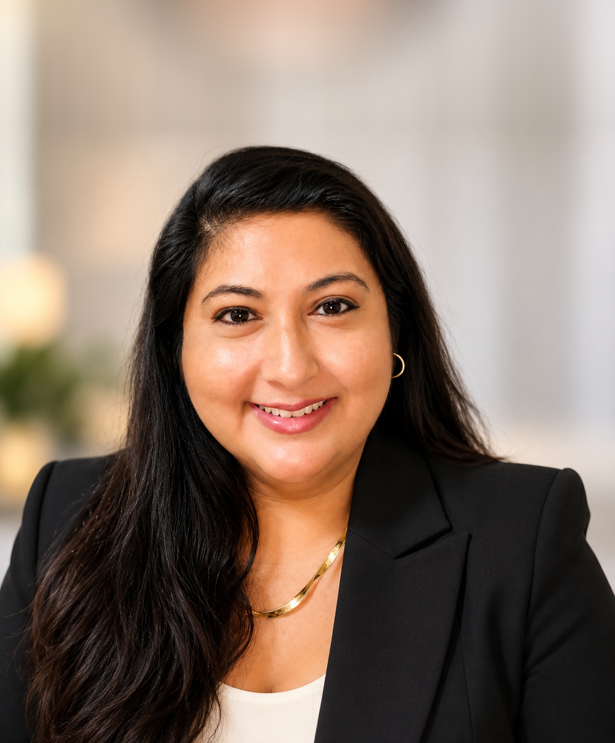 Andrea J. Lopez, founder of Andrea J. Lopez | Digital Atelier, smiling in a tailored black blazer and gold necklace against a softly blurred warm interior
